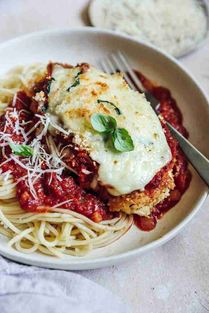 A plated serving of Chicken Parmesan on a bed of spaghetti topped with marinara sauce. The chicken is covered in melted mozzarella and garnished with fresh basil leaves. Grated Parmesan cheese is sprinkled over the spaghetti, and a fork rests beside the dish, ready to enjoy.