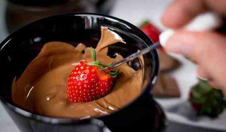 A rich and decadent scene featuring a ripe, juicy strawberry being dipped into a velvety smooth chocolate fondue. The strawberry is held by a fondue fork, poised above a black fondue pot filled with melted chocolate. The chocolate has a glossy sheen, reflecting the light, and contrasts beautifully with the bright red of the strawberry. In the background, blurred elements suggest a cozy atmosphere, perfect for indulging in this sweet treat.