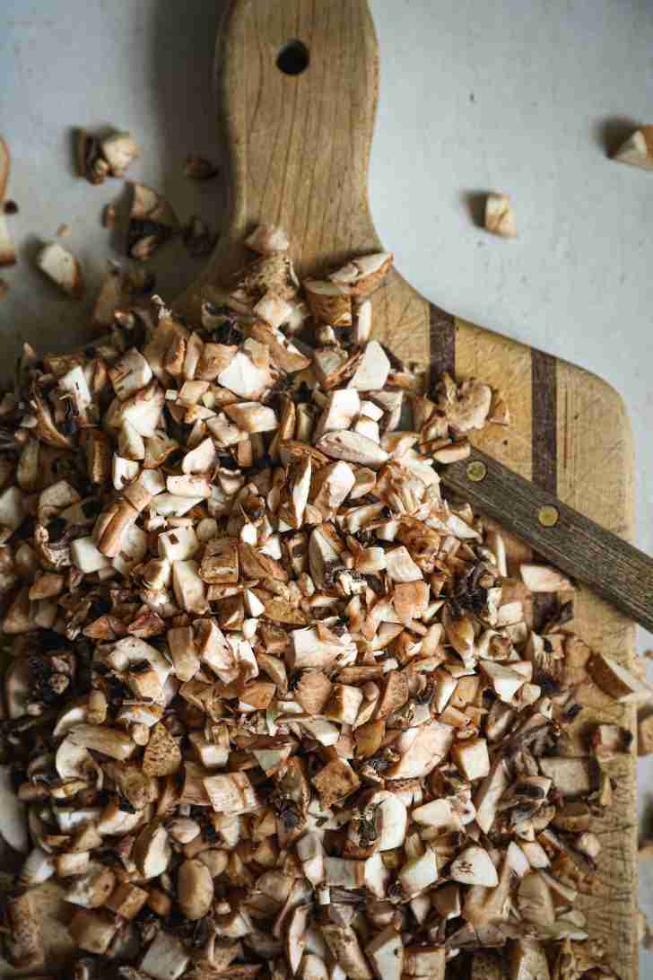 Overhead view of finely chopped mushrooms on a wooden cutting board. The mushrooms are cut into small pieces, ready to be sautéed for the mushroom rolls recipe. Some pieces have fallen off the board, adding a rustic feel to the preparation scene.