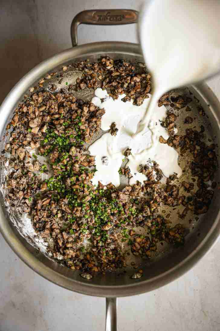 Overhead view of a skillet with sautéed chopped mushrooms and minced chives. Heavy cream is being poured into the skillet, starting to blend with the mushroom mixture to create a creamy filling for the mushroom rolls. The mixture is being cooked and thickened, ready for spreading on the bread slices.