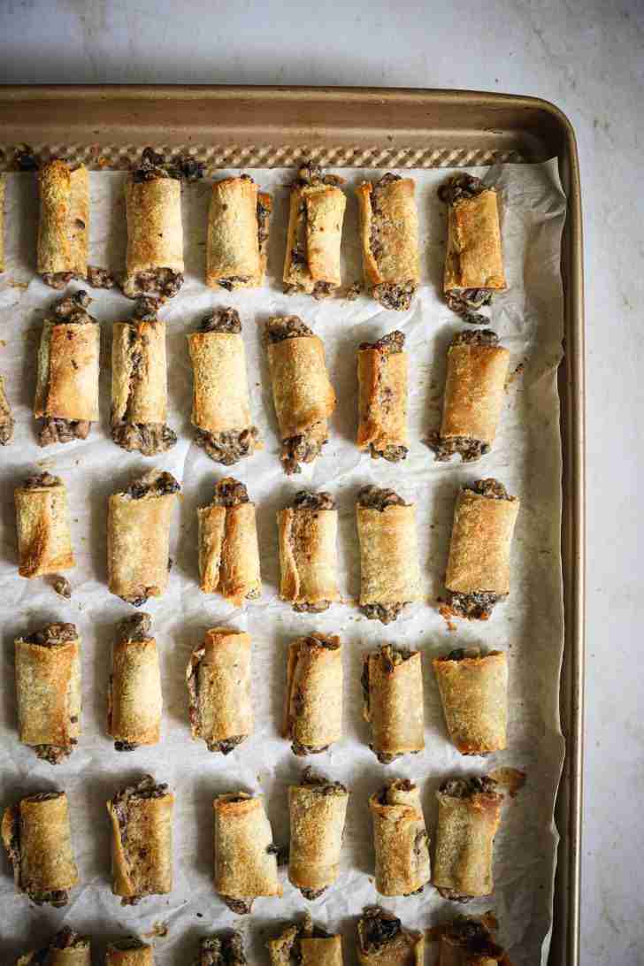 Overhead view of a baking tray lined with parchment paper, filled with golden brown mushroom rolls. The rolls are neatly arranged in rows and have been baked to a crisp, with some of the creamy mushroom filling peeking out from the edges. The rolls are ready to be served as a savory appetizer or snack.