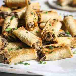 A pile of crispy, golden mushroom rolls garnished with chopped chives, displayed on a white serving dish with a small bowl of herbs in the background.