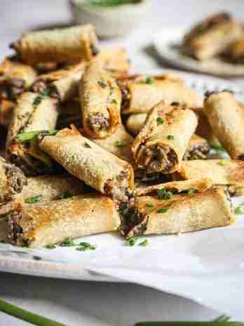 A pile of crispy, golden mushroom rolls garnished with chopped chives, displayed on a white serving dish with a small bowl of herbs in the background.