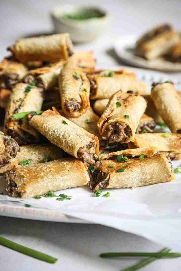 Close-up view of a plate piled with golden brown mushroom rolls, garnished with freshly chopped chives. The rolls are crispy and perfectly toasted, with the savory mushroom filling slightly visible at the ends. A small bowl of extra chives is blurred in the background, adding a touch of freshness to the dish. These mushroom rolls are ready to be enjoyed as a delicious appetizer.