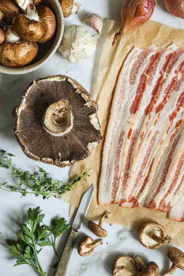 Overhead shot of sliced bacon, assorted mushrooms, garlic, shallots, fresh herbs, and a knife on a kitchen counter, ready for cooking