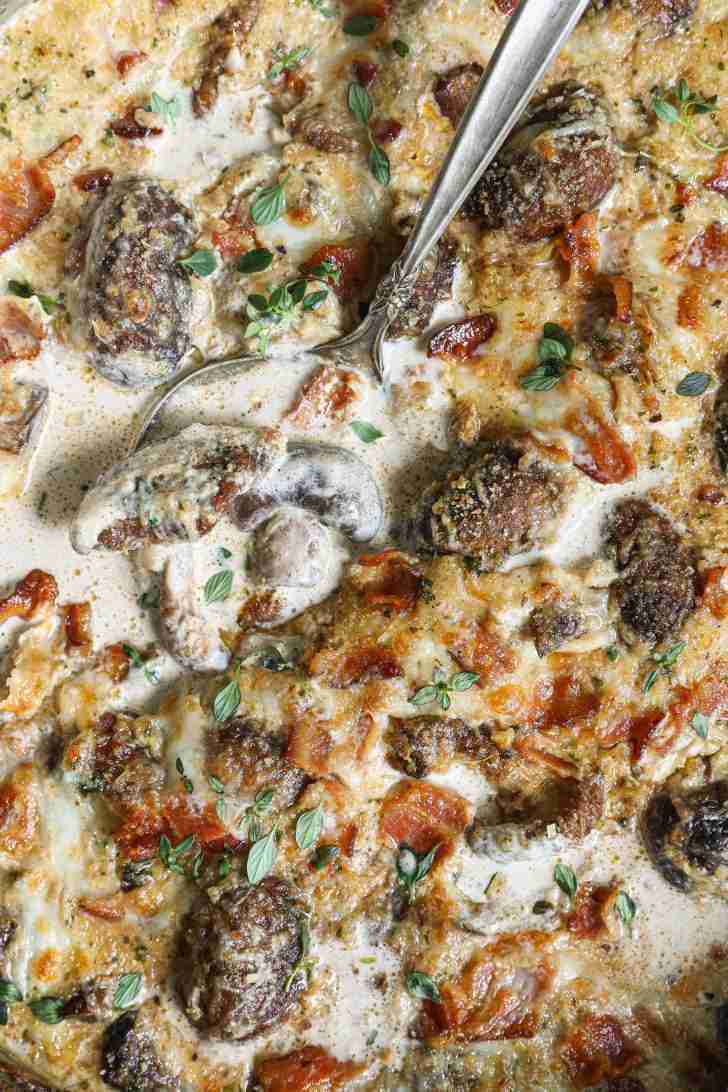 Cheesy mushroom and bacon gratin with a golden, bubbly crust, garnished with fresh herbs, served in a skillet with a spoon for easy serving