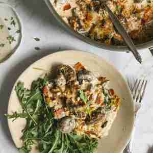 Plate of cheesy mushroom and bacon gratin served with arugula, garnished with toasted pine nuts, next to a skillet of the same dish.