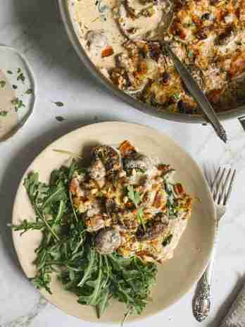 Plate of cheesy mushroom and bacon gratin served with arugula, garnished with toasted pine nuts, next to a skillet of the same dish.