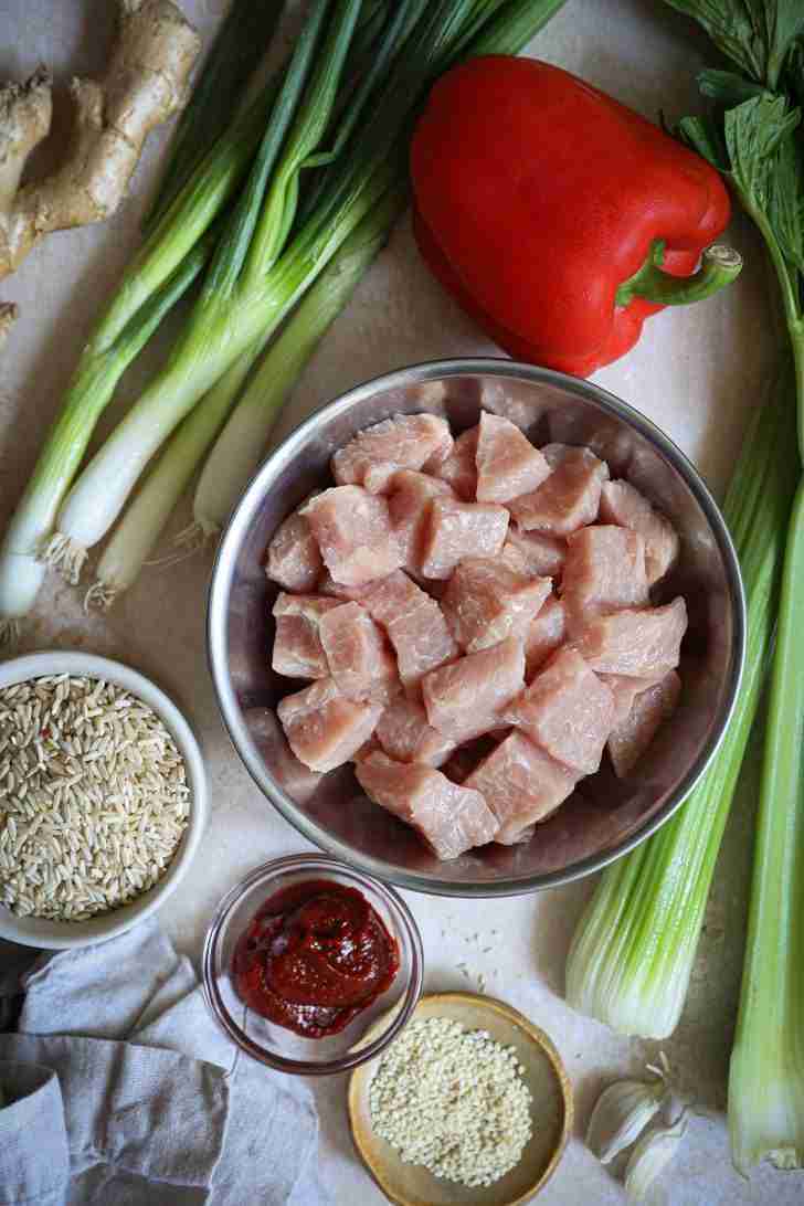 Top-down view of the ingredients for Gochujang Pork Stir-Fry, including diced pork tenderloin in a bowl, red bell pepper, green onions, celery, brown rice, gochujang paste, ginger, and sesame seeds on a light background.