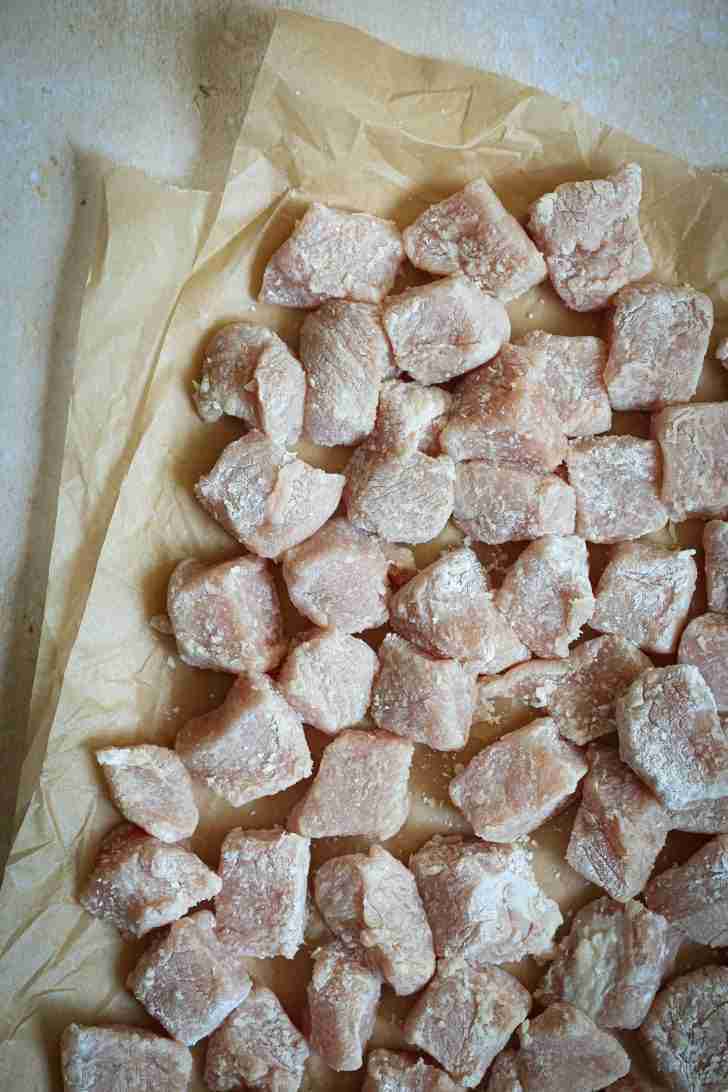 Diced pork tenderloin coated in cornstarch, arranged on parchment paper. The coating creates a light, powdery layer that will crisp up nicely when stir-fried, enhancing texture and flavor.