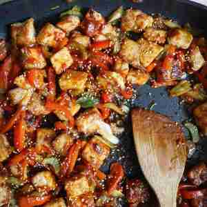 A skillet filled with stir-fried pork, red bell peppers, celery, and green onions coated in a glossy gochujang sauce, topped with sesame seeds.