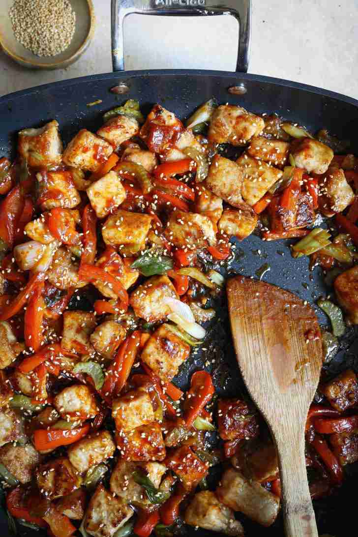 A sizzling wok filled with gochujang-coated pork, bell peppers, and celery, all topped with toasted sesame seeds. The pork pieces are golden and crispy, mingling with vibrant veggies, creating a colorful, mouthwatering stir-fry.