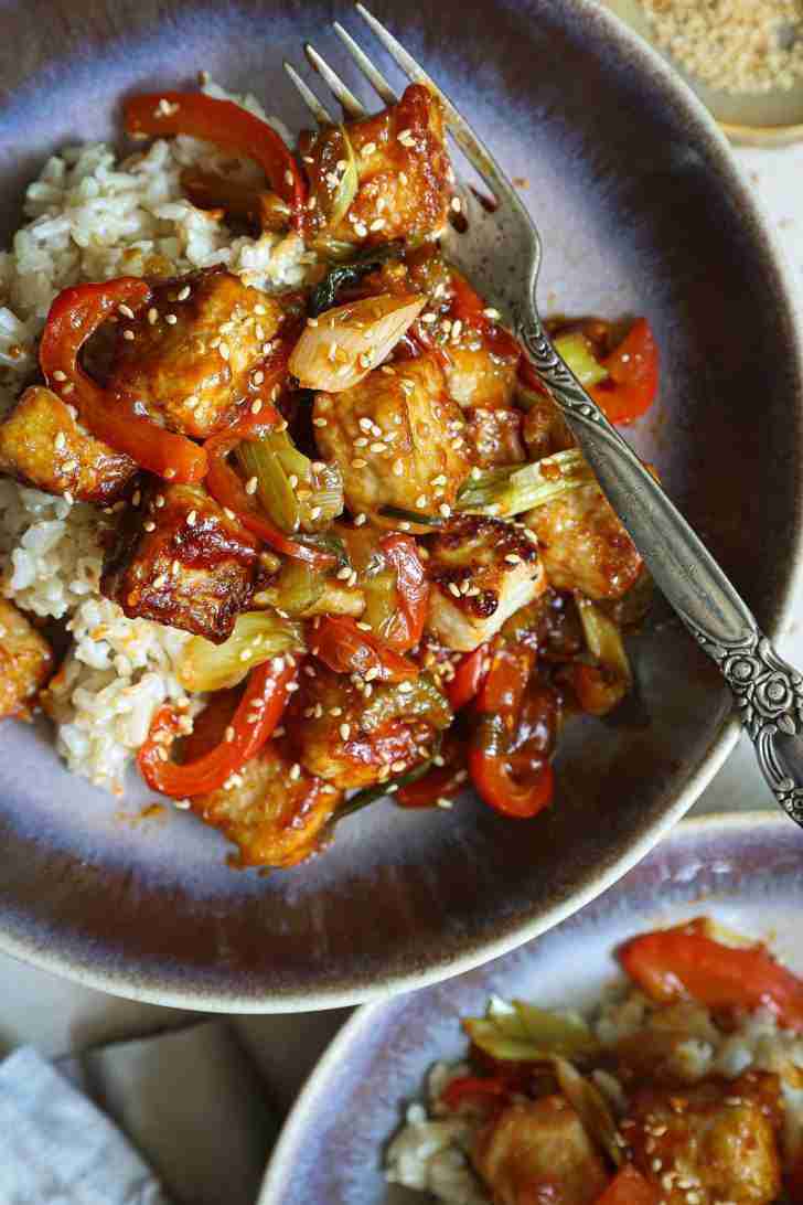 A close-up of a serving of gochujang pork stir-fry over a bed of brown rice, topped with sesame seeds. The pork is crispy, the veggies are vibrant, and the sauce is glossy and rich.