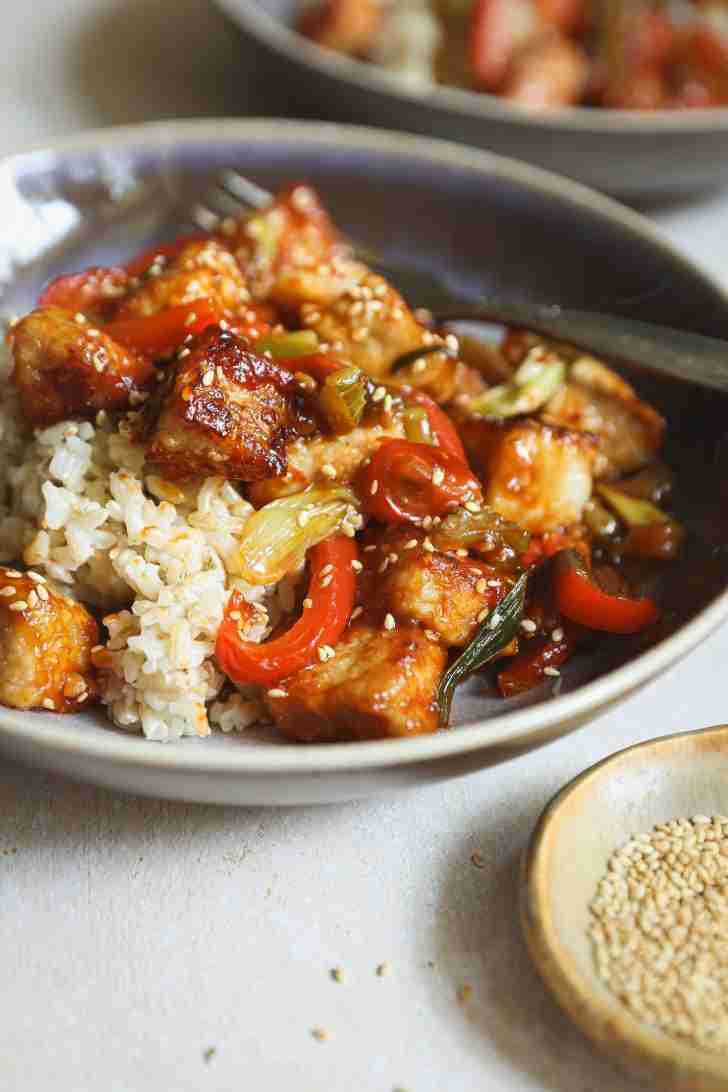 A bowl of gochujang pork stir-fry served over fluffy brown rice, garnished with toasted sesame seeds. The vibrant veggies and glazed pork pieces add a colorful, delicious-looking contrast to the neutral plate.