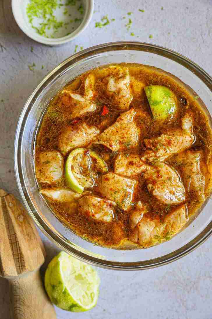 A glass bowl filled with small, bite-sized pieces of chicken marinating in a flavorful liquid. The marinade is a rich, golden-brown color, infused with visible spices and small chili flakes. Lime wedges are mixed in, adding a fresh citrus note. The scene includes a wooden citrus juicer and a squeezed lime half nearby, suggesting the freshly squeezed lime juice used in the marinade. The combination of colors and textures hints at a dish that’s bursting with fresh, zesty flavors.