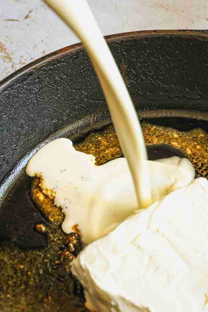 A close-up of heavy cream being poured into a hot skillet, where a block of cream cheese is starting to melt. The skillet is seasoned, with bits of garlic or seasoning visible in the background, adding to the richness of the sauce. The creamy ingredients are beginning to blend, creating the base for a smooth and indulgent sauce. The scene captures the beginning stages of transforming these ingredients into something delicious.