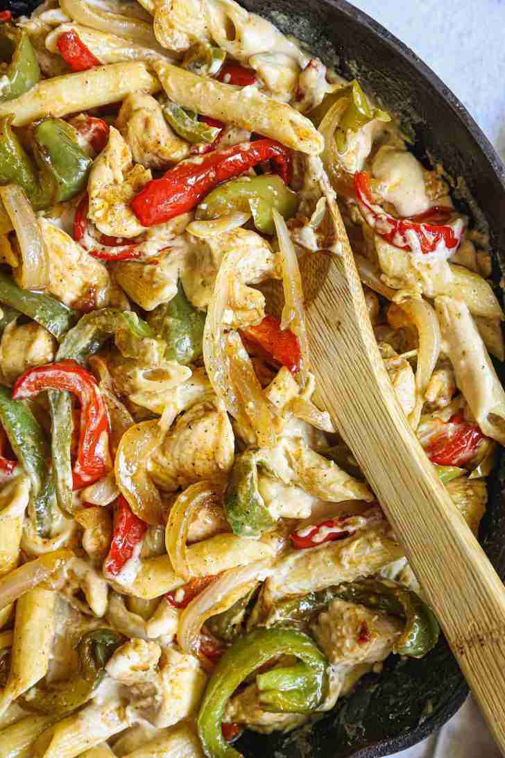 A skillet filled with creamy chicken fajita pasta, featuring tender pieces of chicken, sautéed red and green bell peppers, and onions. The pasta is coated in a rich, cheesy sauce, with a few browned edges where the sauce has caramelized. A wooden spoon rests in the skillet, ready for stirring or serving. The vibrant colors of the vegetables pop against the creamy sauce, making the dish look hearty and flavorful.