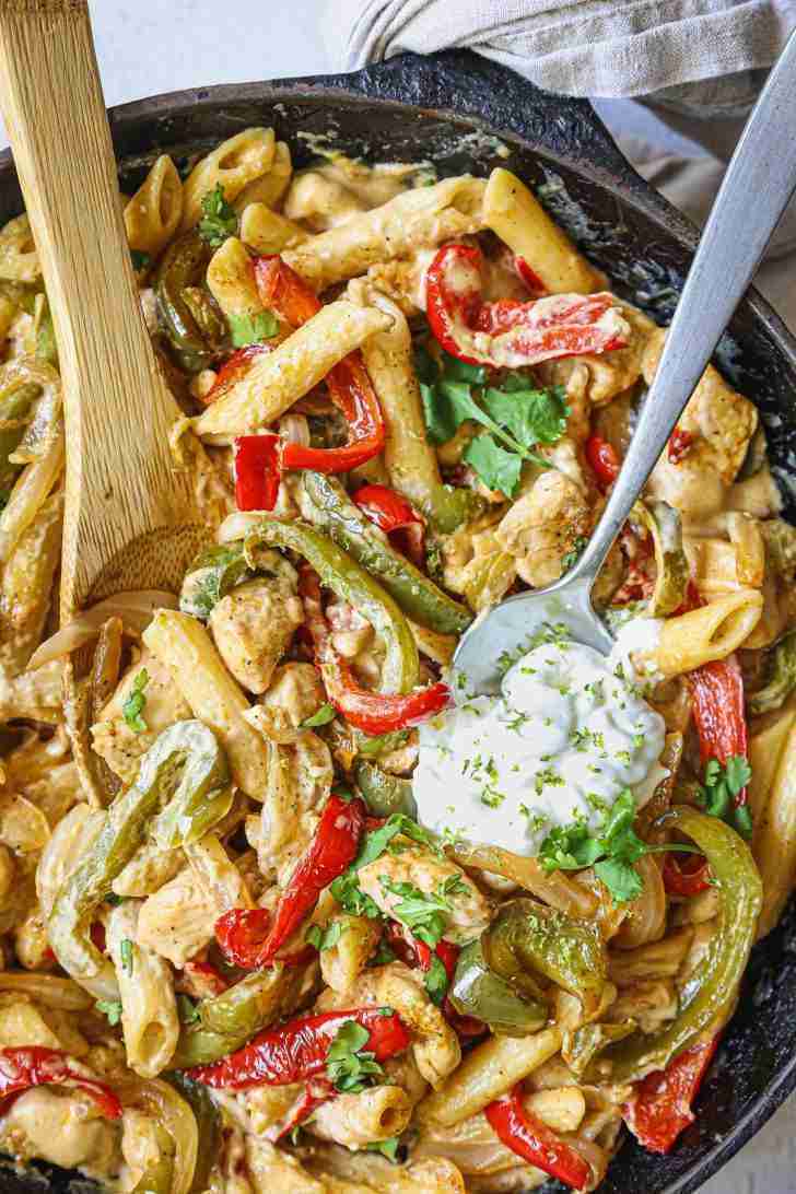 A skillet filled with creamy chicken fajita pasta, topped with sautéed red and green bell peppers, onions, and garnished with fresh cilantro. A dollop of sour cream sits on top, sprinkled with lime zest, adding a fresh and tangy touch. A wooden spoon and a metal spoon are nestled in the pasta, ready for serving. The vibrant colors of the peppers and cilantro contrast beautifully with the creamy sauce, making the dish look both hearty and inviting.