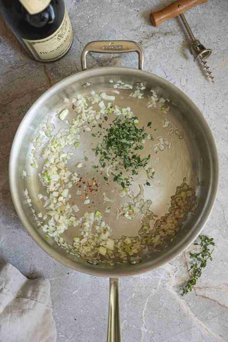 A stainless steel skillet with finely chopped shallots, red pepper flakes, and fresh herbs sautéing in olive oil. A bottle of red wine and a corkscrew are placed nearby on a stone countertop