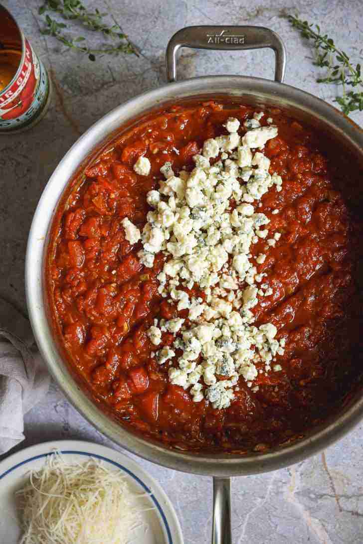 A skillet filled with rich tomato sauce topped with crumbled blue cheese. A small plate of grated Parmesan cheese is placed nearby on a stone countertop, with fresh herbs and a can of diced tomatoes visible in the background.