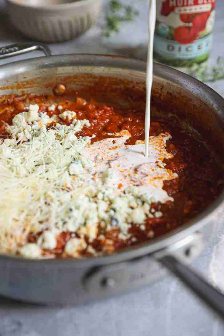 Heavy cream being poured into a skillet filled with a rich tomato sauce, crumbled blue cheese, and grated Parmesan, creating a creamy and cheesy mixture. A can of diced tomatoes and fresh herbs are slightly blurred in the background.