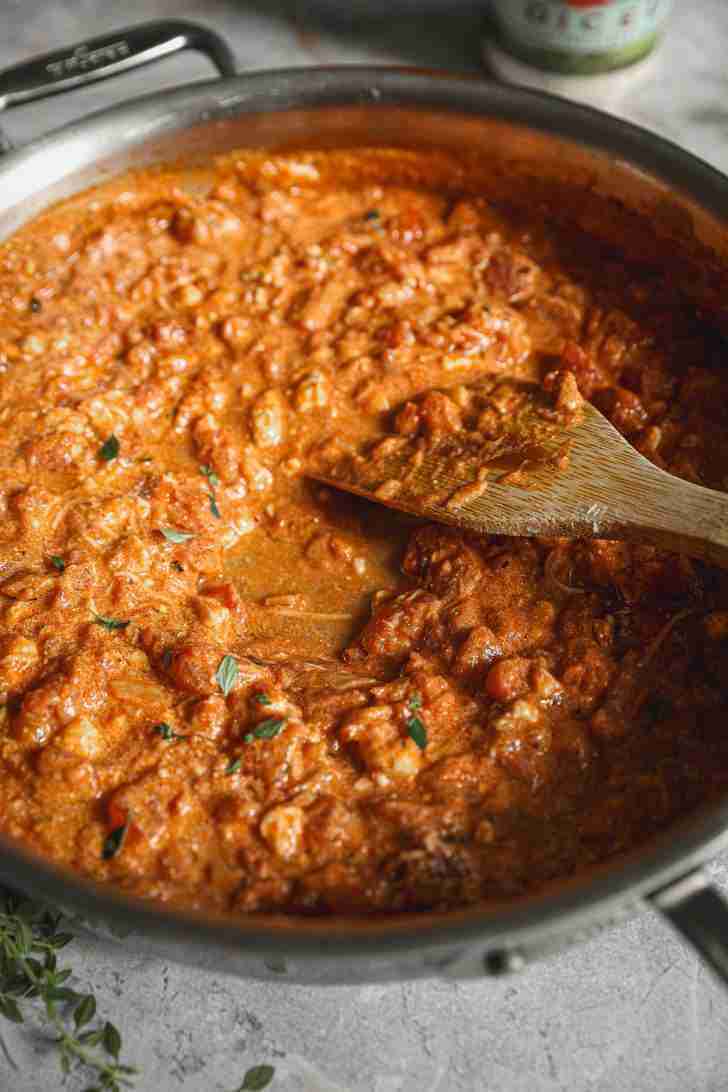 A skillet filled with a rich, creamy tomato and blue cheese sauce being stirred with a wooden spoon. The sauce has a thick, hearty texture with visible chunks of tomatoes and herbs, giving it a rustic appearance.