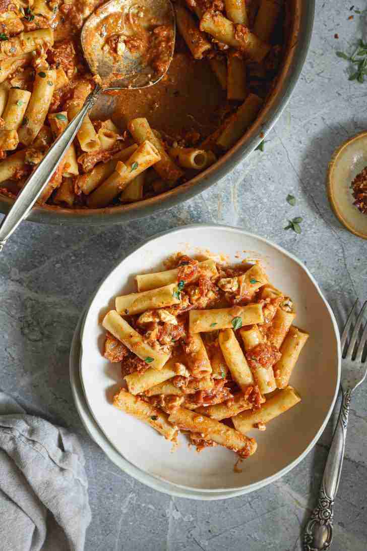 A bowl of ziti pasta in tomato and blue cheese sauce garnished with fresh herbs. Nearby, a skillet with remaining pasta and a serving spoon, with a fork and pepper flakes on the side