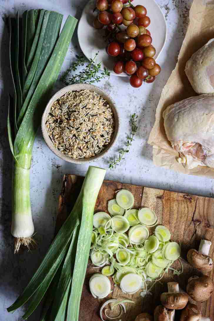 An overhead shot of the ingredients for Harvest Chicken: whole leeks, a bowl of harvest rice medley, a bunch of red seedless grapes, raw chicken pieces, fresh thyme, and sliced cremini mushrooms on a wooden cutting board