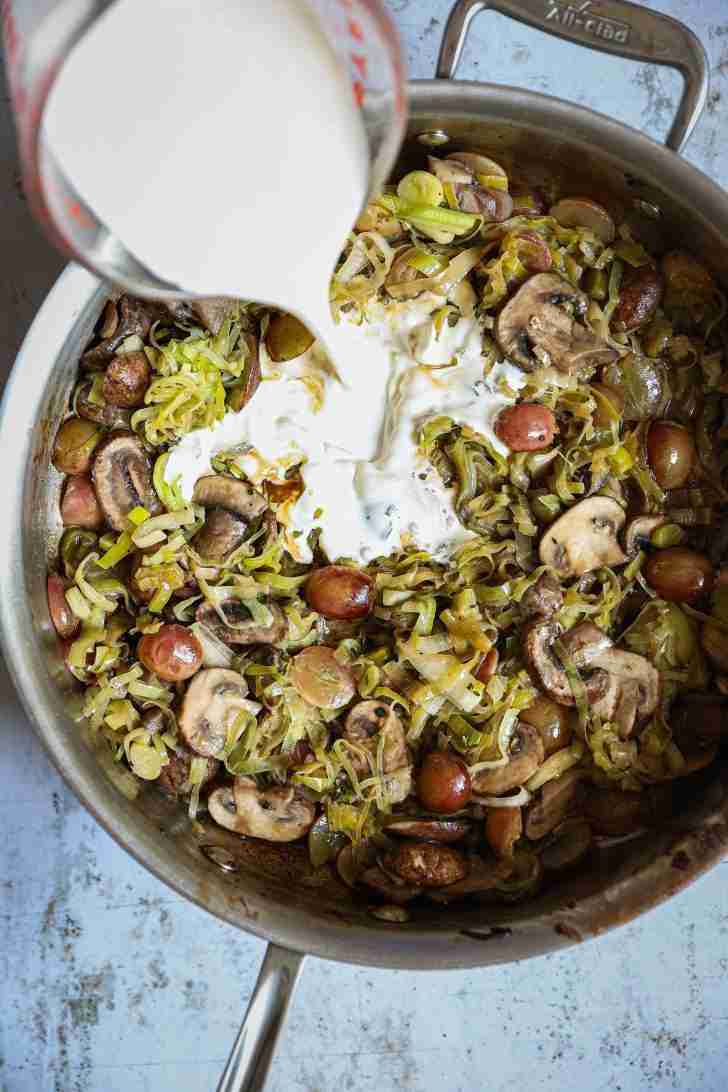 A skillet filled with sautéed leeks, mushrooms, and halved red grapes, as heavy cream is being poured in from a measuring cup, creating a creamy sauce.