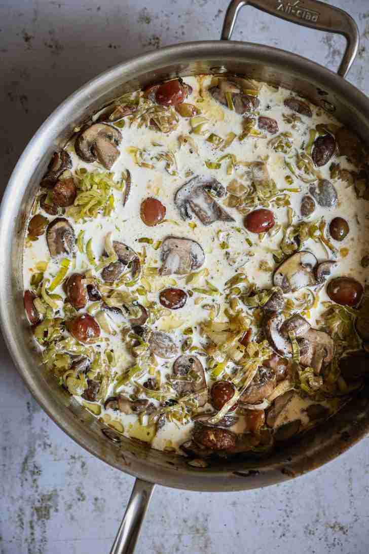 A skillet filled with a creamy mixture of sautéed leeks, mushrooms, and red grapes, all evenly coated in a rich cream sauce, ready to be baked with the chicken