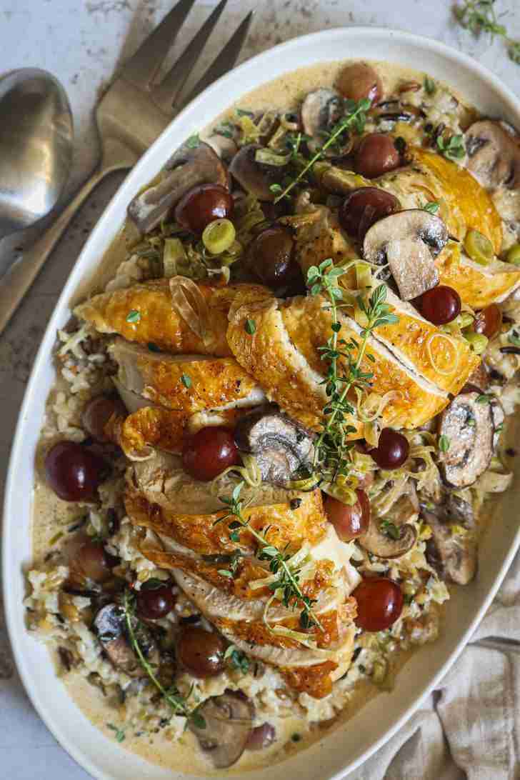 A platter of sliced roasted chicken with sautéed mushrooms, leeks, and grapes, garnished with fresh thyme, served over a bed of harvest rice medley with creamy sauce.