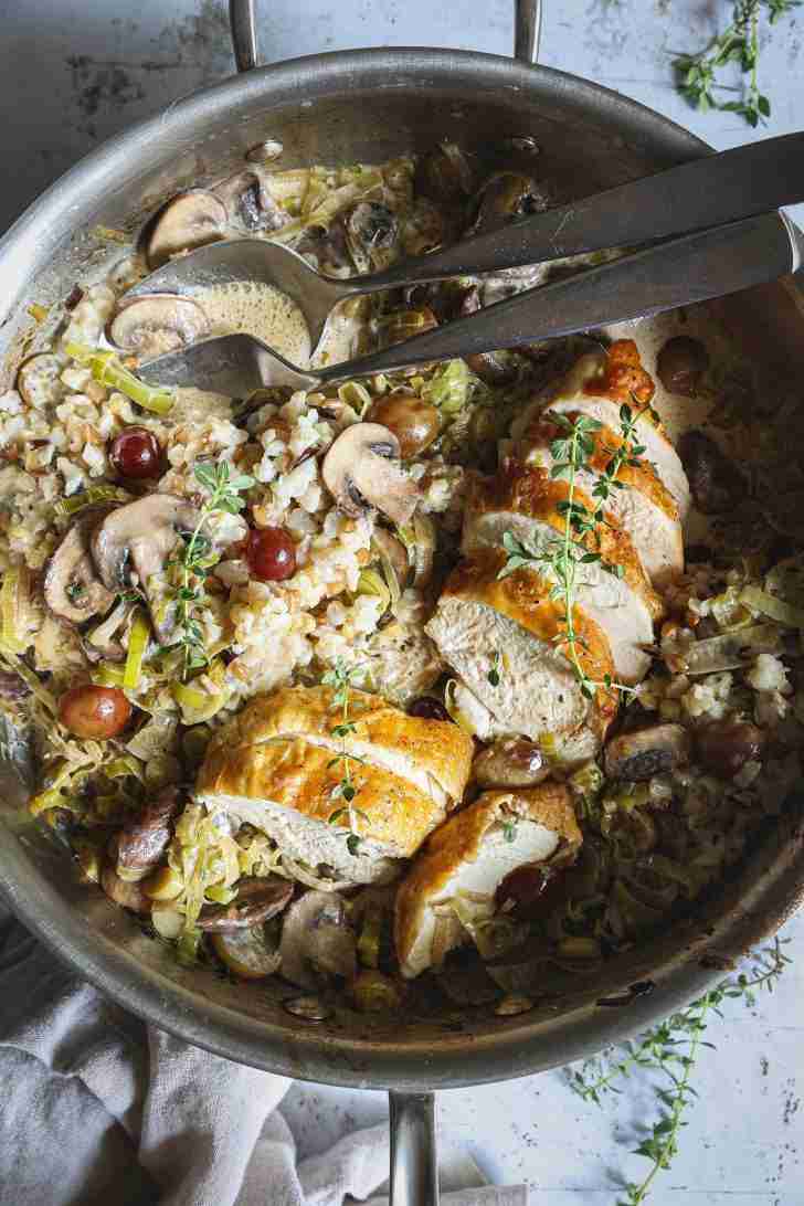 A skillet with sliced roasted chicken, sautéed mushrooms, leeks, and grapes, served over a creamy harvest rice medley and garnished with fresh thyme.