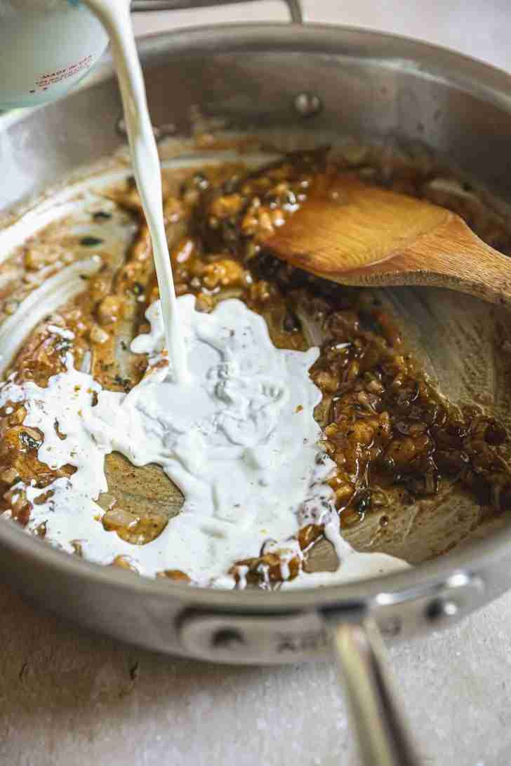 Half-and-half being poured into a skillet with sautéed onions and brandy, forming a creamy, rich base for mac and cheese sauce.