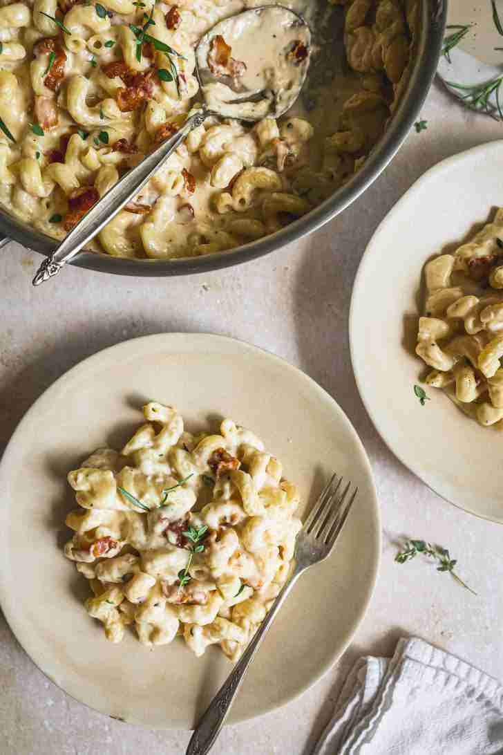 Plated servings of creamy bacon brandy mac and cheese, garnished with fresh herbs, next to the skillet with a serving spoon