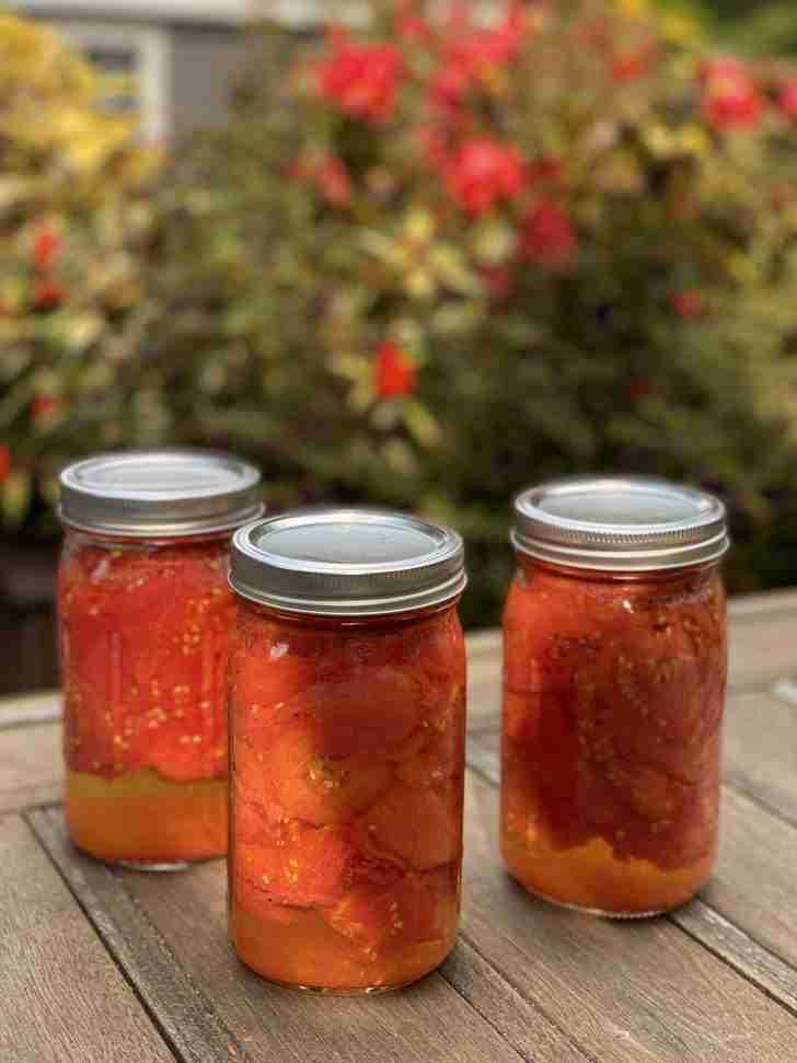 Three jars of freshly canned tomatoes sit on a wooden table outdoors, filled with bright red tomatoes packed tightly in their own juices. The background shows a blurred garden with red and yellow flowers, providing a colorful backdrop to the jars.