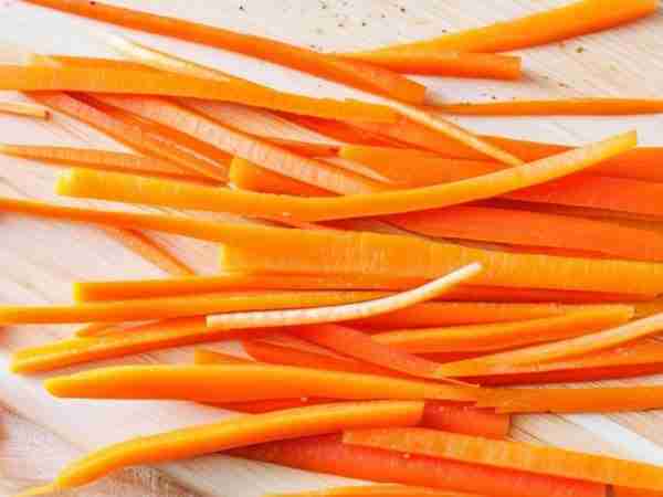 Julienned carrot strips neatly arranged on a rustic, well-worn wooden cutting board with visible grain and scratches, highlighting their bright orange color and precise cuts.