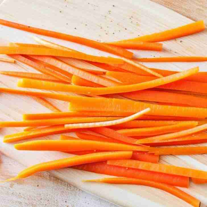 Julienned carrot strips neatly arranged on a rustic, well-worn wooden cutting board with visible grain and scratches, highlighting their bright orange color and precise cuts.