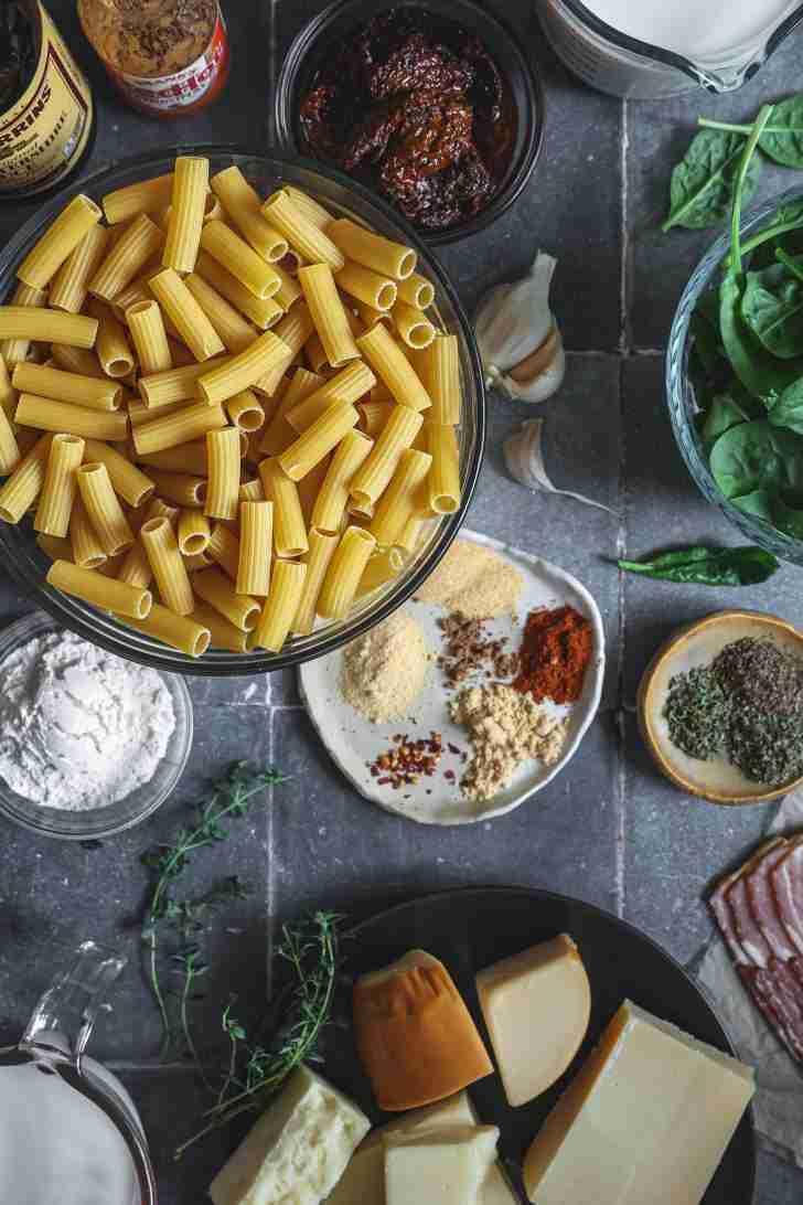 Overhead shot of ingredients for savory mac and cheese: uncooked rigatoni pasta, spices, garlic, spinach, sundried tomatoes, cheeses, flour, and fresh herbs