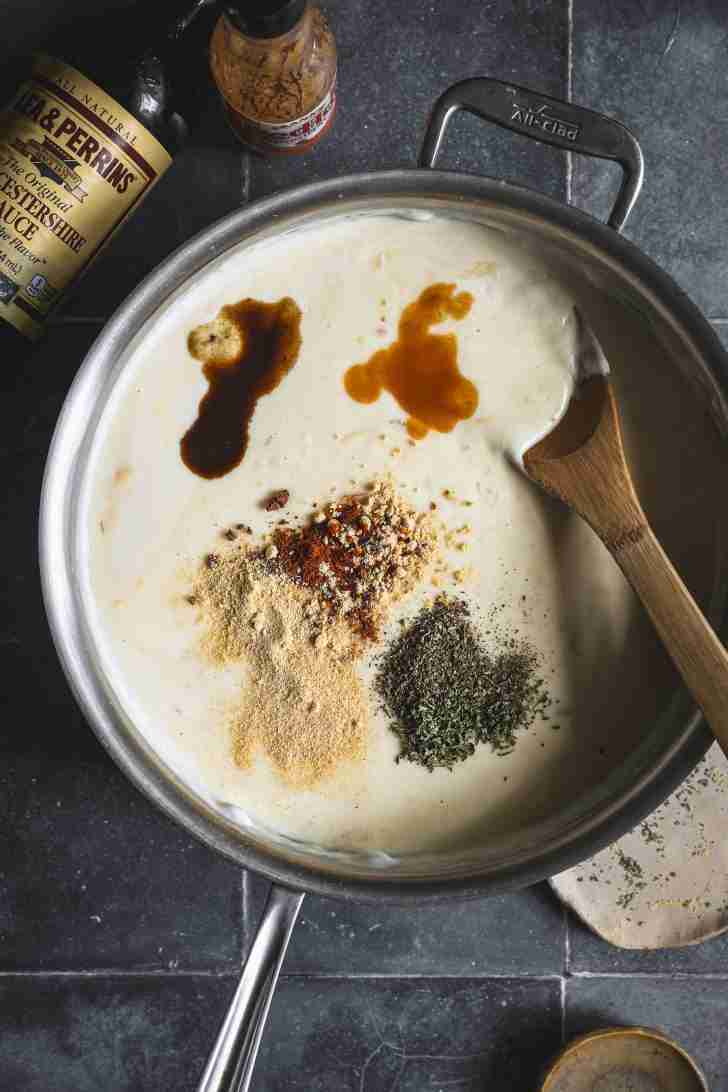 A saucepan with creamy cheese sauce being seasoned with Worcestershire sauce, hot sauce, and various dried spices like mustard powder, paprika, and herbs.