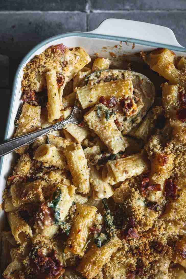 Baked mac and cheese in a casserole dish with golden breadcrumbs on top, crispy bacon, and rigatoni pasta covered in creamy cheese sauce.