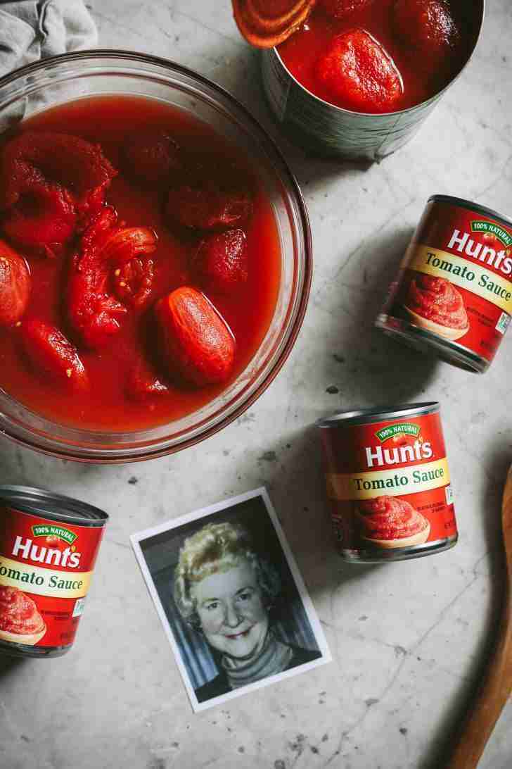 A bowl filled with whole tomatoes in their juice on a marble countertop next to three cans of Hunt's tomato sauce. A black-and-white photo of my smiling mom adds a warm, personal touch to the scene.