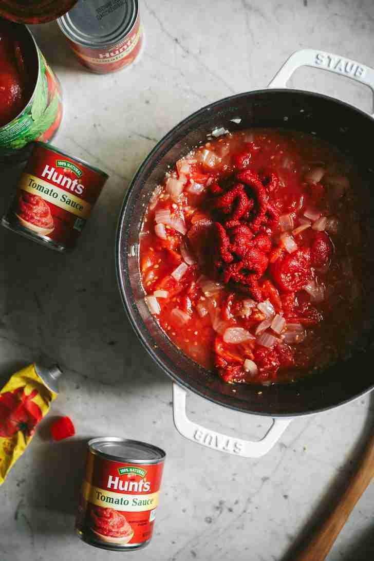 An overhead view of a black Staub pot filled with a mix of sautéed onions, whole tomatoes, and a generous swirl of tomato paste, showing the base for a hearty spaghetti sauce. Hunts tomato sauce cans.