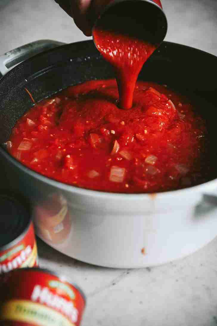 A close-up shot of rich, red tomato sauce being poured from a can into a white pot filled with simmering spaghetti sauce. The sauce thickens with each addition, blending perfectly with sautéed onions and crushed tomatoes. In the background, extra cans of Hunts tomato sauce are slightly blurred, ready to enhance the dish further.