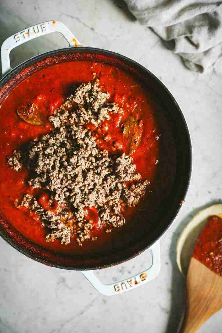 A white Staub pot filled with a simmering, rich red tomato sauce, topped with freshly browned ground beef. Bay leaves peek through the surface, infusing the sauce with their aromatic flavor. A wooden spoon rests nearby, slightly smeared with sauce, ready to stir and blend all the ingredients together into a hearty, delicious dish.