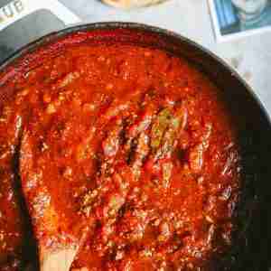 A bubbling pot of rich, homemade tomato sauce, loaded with ground beef and seasoned to perfection with bay leaves and herbs. A wooden spoon is halfway submerged, coated with the deep red sauce. In the background, a can of Hunt's Tomato Sauce and a photo of my smiling mom add a touch of nostalgia to the cooking scene, making it feel warm and personal.