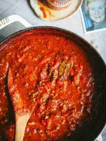 A bubbling pot of rich, homemade tomato sauce, loaded with ground beef and seasoned to perfection with bay leaves and herbs. A wooden spoon is halfway submerged, coated with the deep red sauce. In the background, a can of Hunt's Tomato Sauce and a photo of my smiling mom add a touch of nostalgia to the cooking scene, making it feel warm and personal.