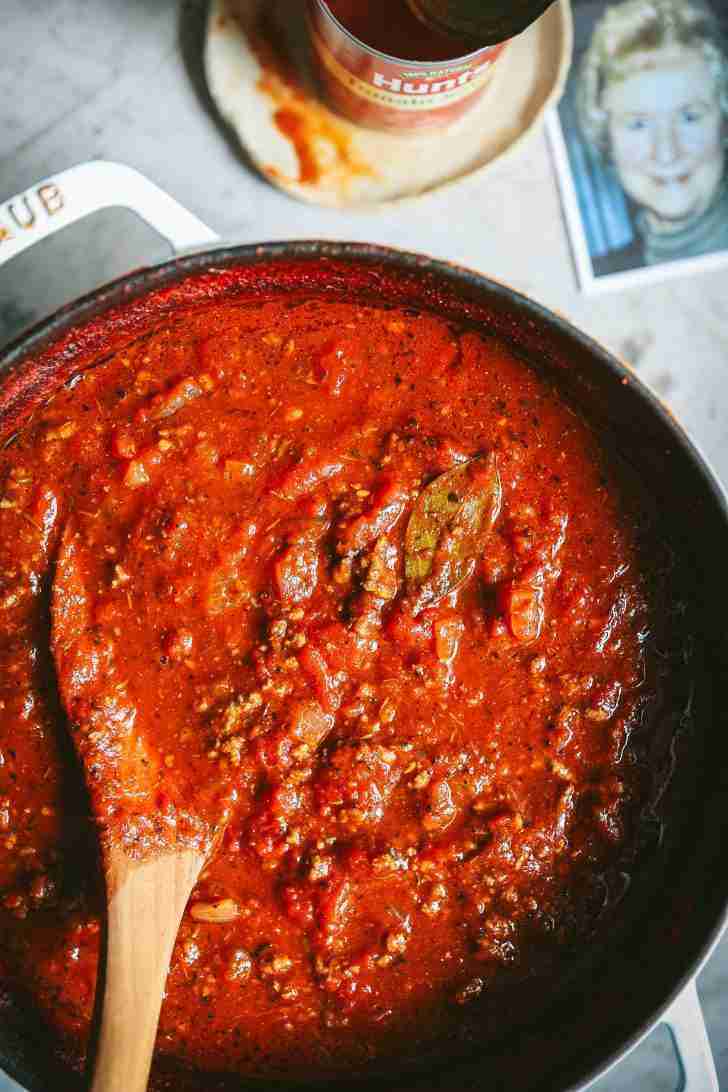 A bubbling pot of rich, homemade tomato sauce, loaded with ground beef and seasoned to perfection with bay leaves and herbs. A wooden spoon is halfway submerged, coated with the deep red sauce. In the background, a can of Hunt's Tomato Sauce and a photo of my smiling mom add a touch of nostalgia to the cooking scene, making it feel warm and personal.