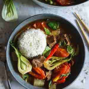 A bowl of beef sinigang served with a mound of steamed rice, featuring vibrant bok choy, tender beef chunks, tomatoes, and a red chili pepper.