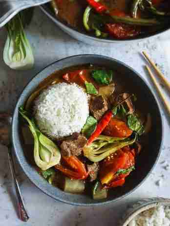 A bowl of beef sinigang served with a mound of steamed rice, featuring vibrant bok choy, tender beef chunks, tomatoes, and a red chili pepper.