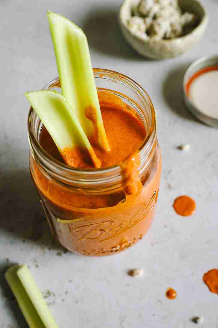 A jar filled with Blue Heat Wing Sauce has celery sticks inserted, with sauce drips on the side of the jar and some splatters on the countertop.